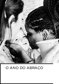 Fotografia em preto e branco. Mulher beijando bebê enquanto outra mulher segurando copo observa de costas. Abaixo “O ANO DO ABRAÇO”.