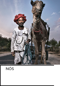 Fotografia. Cuidador de camelo em Rajastão, Índia, usa roupas brancas e turbante vermelho. Posa ao lado de seu camelo.  