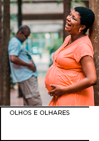 Fotografia. No primeiro plano, uma mulher grávida enquanto sorri acaricia a barriga. No segundo plano, um homem repete o gesto.