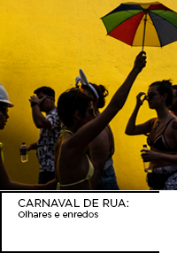 Fotografia. Em um fundo amarelo, pessoas comemoram o bloco de carnaval em Vitória. Uma delas levanta um pequeno guarda-chuva colorido.