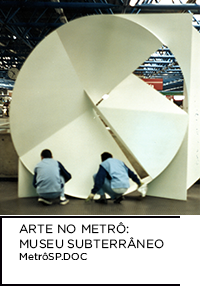 Fotografia. Homens colocando escultura “A Roda” de Emanoel Araújo. Abaixo “ARTE NO METRÔ: MUSEU SUBTERRÂNEO MetrôSP.Doc”.