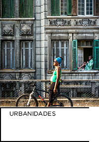 Fotografia. Mulher ao lado de bicicleta em frente a um prédio. Abaixo “Urbanidades”.